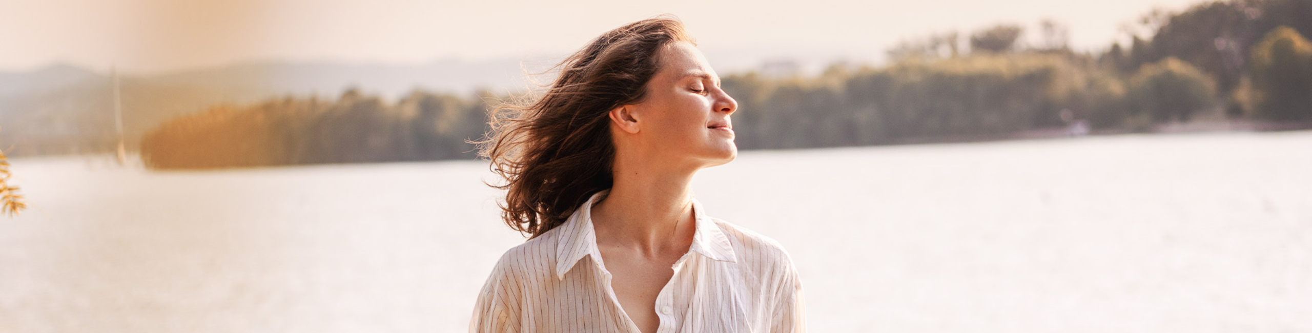 Žena stoji napolju pored jezera uživajući u suncu zatvorenih očiju